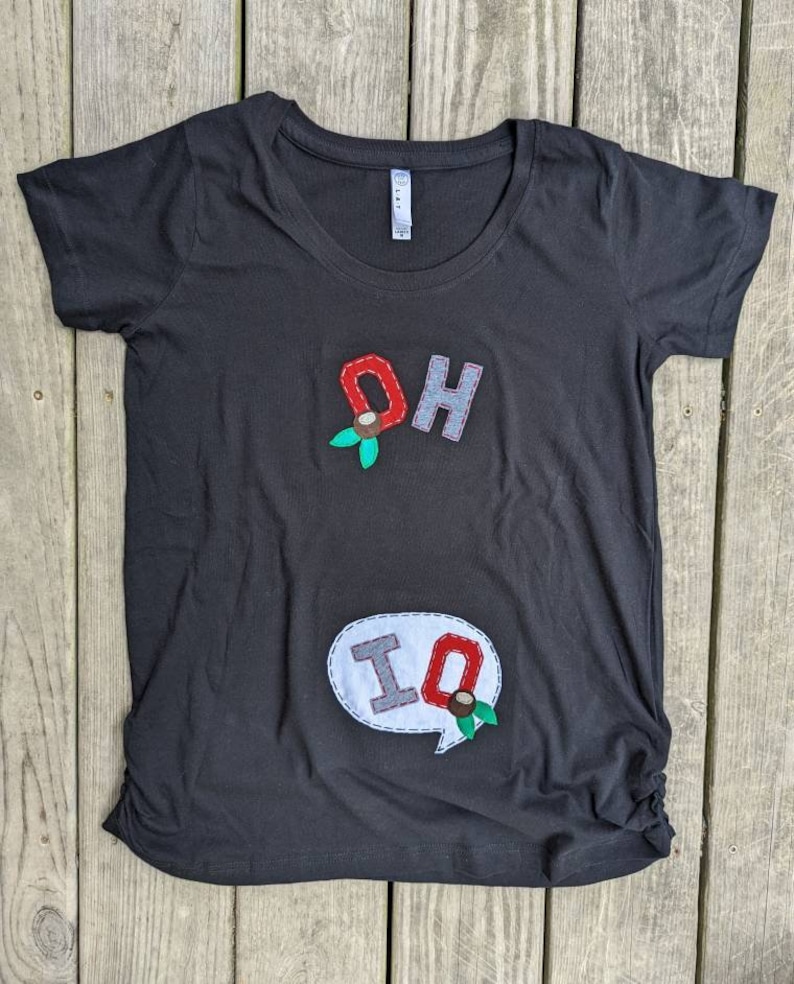 Pode incluir: Camiseta preta com um "OH" vermelho e cinza e um bal&atilde;o de fala branco com um "IQ" cinza em costura vermelha. As letras s&atilde;o decoradas com folhas verdes e bagas vermelhas.