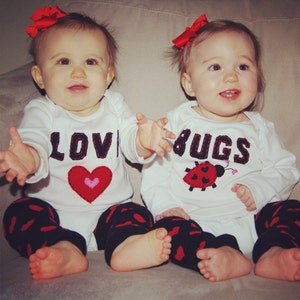 May include: Two babies wearing white long-sleeved shirts with red bows in their hair. One shirt says "LOVE" with a red heart and the other says "BUGS" with a red ladybug. Both babies are wearing black and red leggings.