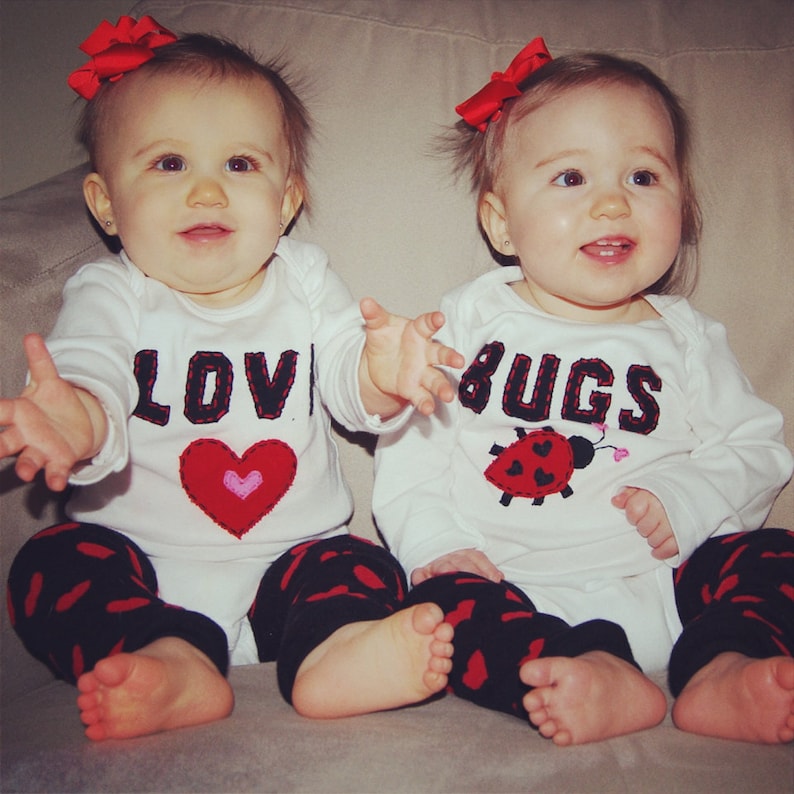 May include: Two babies wearing white long-sleeved shirts with red bows in their hair. One shirt says "LOVE" with a red heart and the other says "BUGS" with a red ladybug. Both babies are wearing black and red leggings.