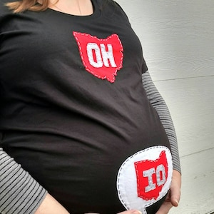 Pode incluir: Uma camiseta preta com um contorno de feltro vermelho do estado de Ohio com as letras "OH" em feltro branco. Um segundo contorno de feltro vermelho do estado de Ohio com as letras "IO" em feltro branco est&aacute; na barriga da camiseta.