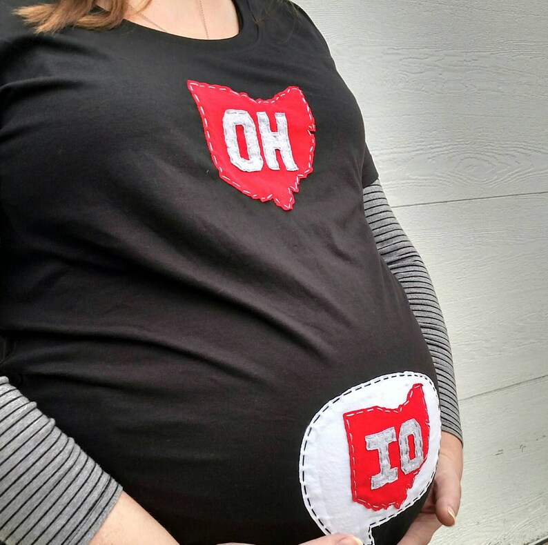 Pode incluir: Uma camiseta preta com um contorno de feltro vermelho do estado de Ohio com as letras "OH" em feltro branco. Um segundo contorno de feltro vermelho do estado de Ohio com as letras "IO" em feltro branco est&aacute; na barriga da camiseta.