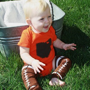 May include: A baby wearing an orange bodysuit with a brown Ohio state outline on the front. The baby is wearing brown and white football leg warmers and is sitting in a metal tub.