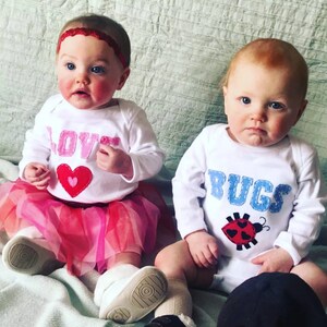 May include: Two babies wearing white onesies with embroidered designs. The baby on the left is wearing a pink tutu and has a red headband. The onesie says "LOVE" with a red heart. The baby on the right is wearing a white onesie that says "BUGS" with a red ladybug.