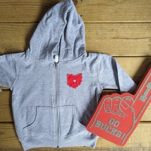 May include: A grey hooded sweatshirt with a red heart-shaped Ohio patch on the front. A red foam finger with the words "Go Bucks!" is in the background.