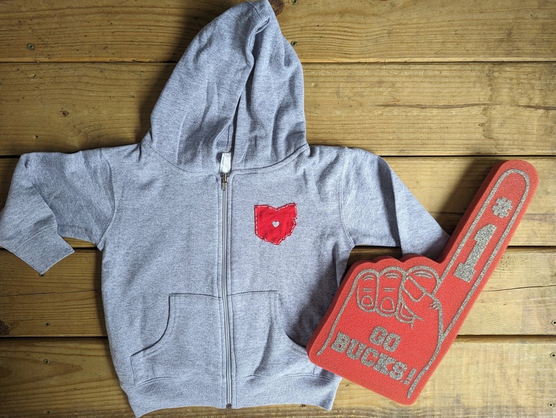 May include: A grey hooded sweatshirt with a red heart-shaped Ohio patch on the front. A red foam finger with the words "Go Bucks!" is in the background.