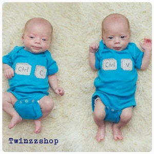 Op de afbeelding: Twee baby's dragen blauwe rompers met witte plekken waarop "Ctrl C" en "Ctrl V" staat. De baby's liggen op een wit, pluche oppervlak.
