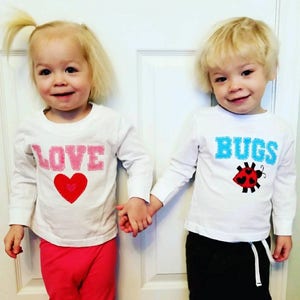 May include: Two children wearing white long-sleeved shirts with embroidered designs. The girl's shirt says "LOVE" with a pink heart. The boy's shirt says "BUGS" with a red ladybug.