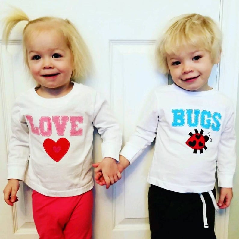May include: Two children wearing white long-sleeved shirts with embroidered designs. The girl's shirt says "LOVE" with a pink heart. The boy's shirt says "BUGS" with a red ladybug.