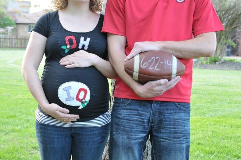 Pode incluir: Uma mulher gr&aacute;vida vestindo uma camiseta preta com as letras "OH" e "IQ" nela. Ela est&aacute; de p&eacute; ao lado de um homem que est&aacute; vestindo uma camiseta vermelha com uma bola de futebol em suas m&atilde;os. A bola de futebol tem a data "6-22-14" escrita nela.