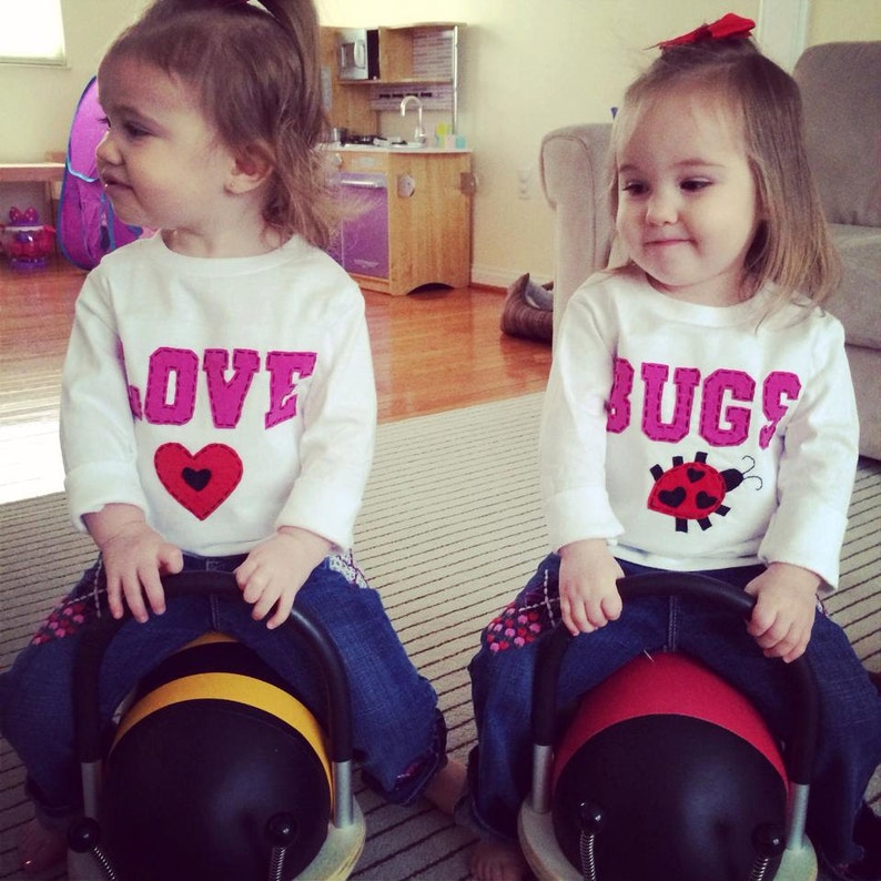 May include: Two young girls wearing white long-sleeved shirts with pink lettering. One shirt says "LOVE" with a pink heart and the other says "BUGS" with a ladybug graphic. Both girls are wearing blue jeans and riding on black and yellow toy vehicles.