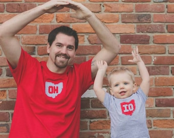 May include: A father and son wearing matching red and grey t-shirts with the state of Ohio outlined in white. The father is holding his hands above his head in an 'O-H' gesture, while the son is standing with his arms raised. The shirts have the letters 'OH' and 'IO' on them.