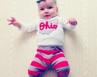 May include: A baby wearing a white onesie with a red "Ohio" graphic on the front and gray and pink striped pants.