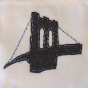 Peut inclure: Une appliqu&eacute; en feutre gris fonc&eacute; repr&eacute;sentant le pont de Brooklyn, avec des coutures bleues qui d&eacute;limitent le pont et ses tours. L'appliqu&eacute; est cousu sur un fond en tissu blanc.