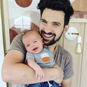 Pode incluir: Um homem com cabelo escuro e barba está segurando um bebê nos braços. O bebê está vestindo um macacão cinza com um desenho de um copo de uísque e a palavra "neat" impressa nele. O homem está sorrindo e olhando para a câmera.