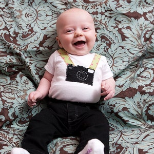 May include: A baby wearing a white onesie with a black camera design and green and pink straps. The onesie has a stitched outline. The baby is wearing black trousers and is lying on a brown and blue patterned fabric.