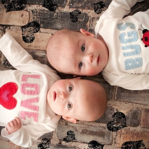 May include: Two babies wearing white onesies with different designs. One onesie has the word "LOVE" in pink and red letters and a red heart. The other onesie has the word "BUGS" in blue letters and a red ladybug. The babies are lying on a brick surface with black hearts scattered around them.
