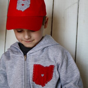 May include: A young person wearing a grey hooded sweatshirt with a red Ohio state outline patch on the front pocket. The sweatshirt has a zip fastening. The person is also wearing a red baseball cap with a grey Ohio state outline patch on the front.