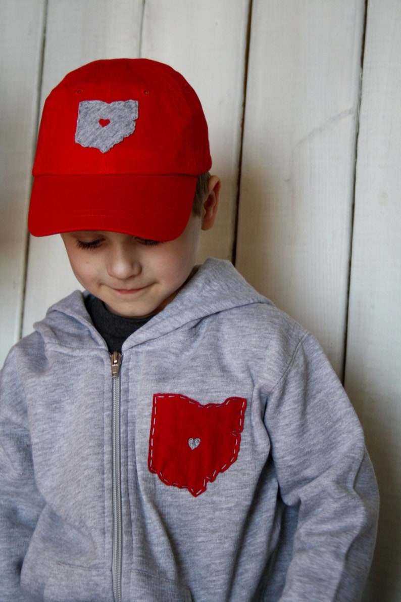 May include: A young person wearing a grey hooded sweatshirt with a red Ohio state outline patch on the front pocket. The sweatshirt has a zip fastening. The person is also wearing a red baseball cap with a grey Ohio state outline patch on the front.