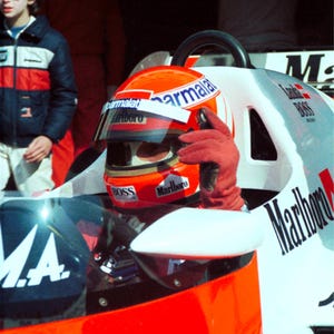May include: A red, white, and blue race car with the Marlboro logo on the side. The driver is wearing a red helmet with a Marlboro logo and a red racing suit.