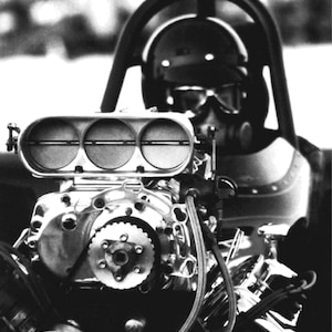 Puede incluir: Una fotografía en blanco y negro de un motor de coche de carreras con un conductor en la cabina. El motor tiene un colector de admisión de cromo grande con tres aberturas redondas. El conductor lleva un casco y gafas.