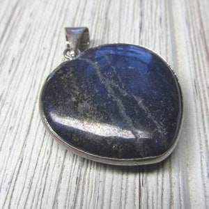Puede incluir: Un colgante de plata en forma de coraz&oacute;n con una piedra azul oscuro y negra. La piedra tiene un patr&oacute;n estriado y un acabado brillante.