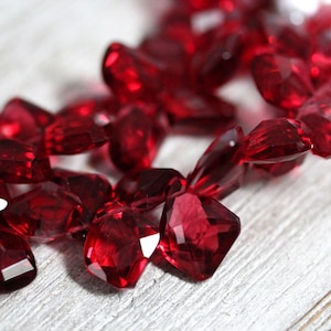 Puede incluir: Una hebra de cuentas de piedras preciosas facetadas de color rojo intenso. Las cuentas están talladas en forma cuadrada con muchas facetas. Las cuentas están enhebradas en un hilo transparente.