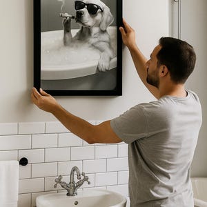 May include: A framed black and white print of a dog in sunglasses, smoking a cigar while relaxing in a bathtub. The print is being hung on a white tiled wall in a bathroom setting.
