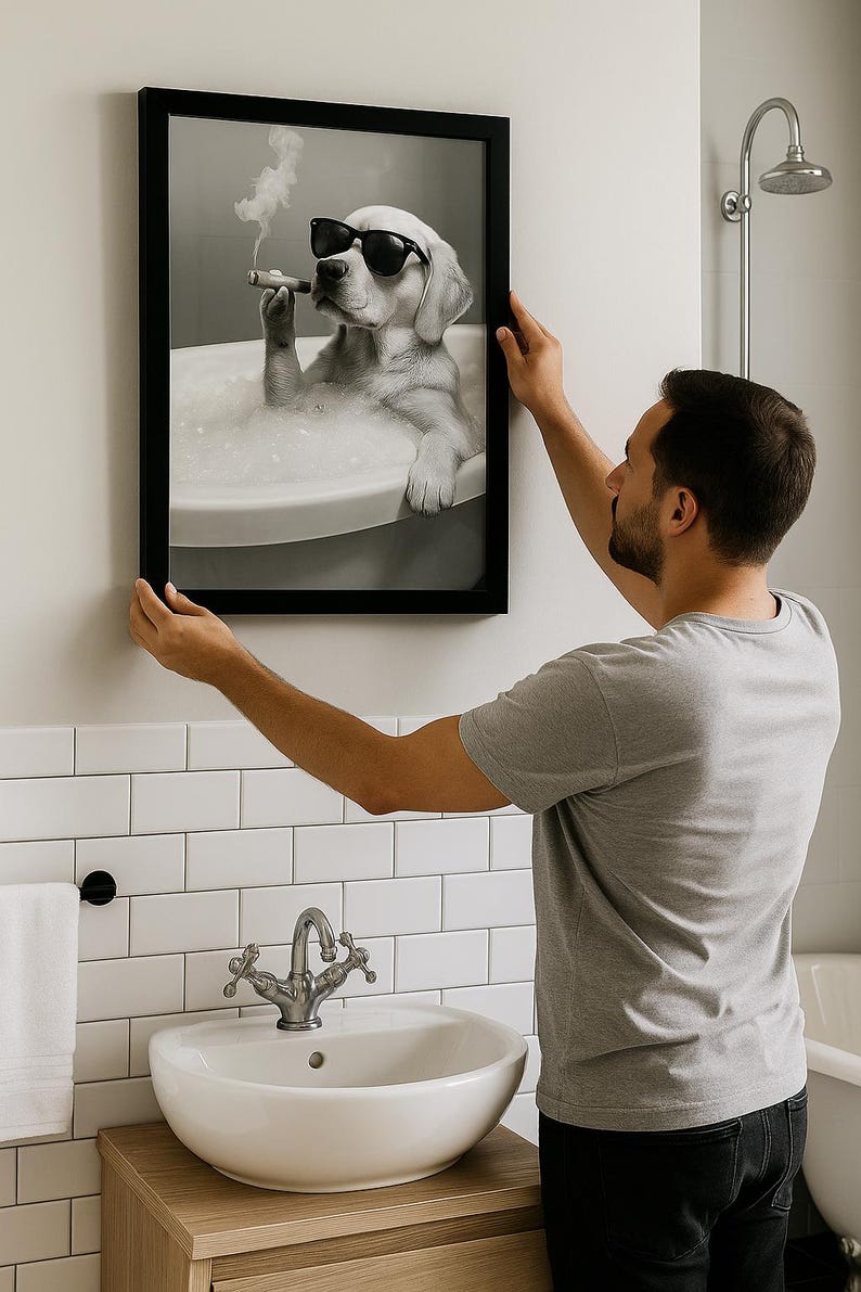 May include: A framed black and white print of a dog in sunglasses, smoking a cigar while relaxing in a bathtub. The print is being hung on a white tiled wall in a bathroom setting.