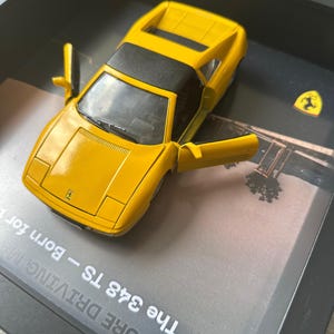 Może przedstawiać: Żółty model samochodu Ferrari 348 TS z otwartymi drzwiami, prezentowany w czarnej ramie. Samochód ma czarny dach i widoczne jest logo Ferrari. Widoczny jest również tekst "PURE DRIVING - Born for".