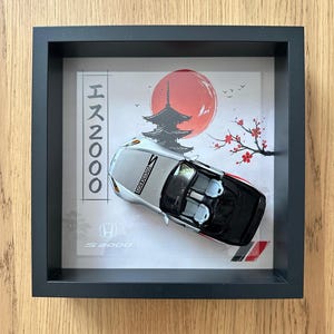 Könnte beinhalten: Gerahmtes Kunstwerk mit einem silbernen und schwarzen Honda S2000 Modellauto. Der Hintergrund zeigt eine japanische Landschaft mit roter Sonne, Pagode und Kirschblüten. Der Rahmen ist schwarz.