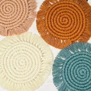May include: Four handmade macrame coasters with fringe. The coasters are in shades of beige, brown, white, and blue.