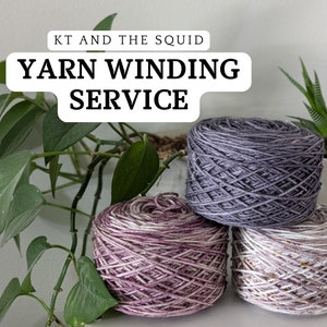 May include: Three skeins of wound yarn in shades of purple, gray, and white are stacked on a white surface. The text "KT AND THE SQUID" and "YARN WINDING SERVICE" are displayed above the yarn. Green plants are in the background.