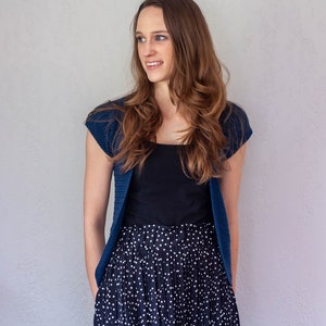 May include: A woman wearing a blue cardigan over a black tank top and a black and white polka dot skirt.
