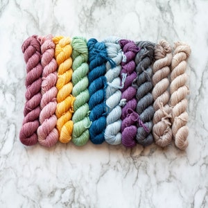 May include: A collection of ten skeins of yarn in a rainbow of colors, including shades of pink, yellow, green, blue, purple, gray, and beige. The yarn is arranged in a row on a white and gray marble surface.