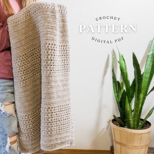 May include: A beige crochet blanket with a textured pattern is held up. The blanket is folded, showing the detailed stitch work. Text on the image reads "CROCHET PATTERN DIGITAL PDF". A potted snake plant is in the background.