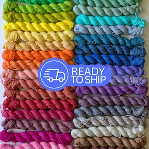 May include: A collection of colorful yarn skeins arranged in rows, showcasing a spectrum of hues from greens and yellows to blues, reds, and browns. The image includes the text "READY TO SHIP" in a blue circle with a truck icon.