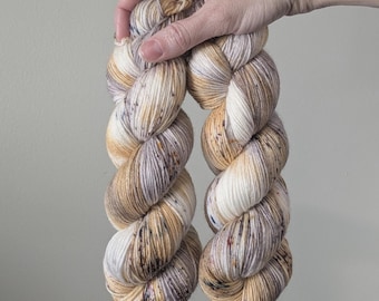 May include: Two skeins of hand-dyed yarn in a neutral color palette. The yarn features a blend of white, tan, and brown hues, creating a speckled effect. The yarn is held up against a neutral background.