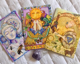 May include: Three colourful cards with illustrations of a Buddha, two women with a sun above them, and a woman playing a drum. The cards have the text "Sisters Under" on the bottom.