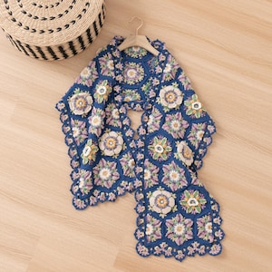 May include: A blue crochet shawl with a floral design. The shawl has a repeating pattern of hexagonal shapes with cream, lavender, and yellow flower motifs. The edges are scalloped, and the shawl is displayed on a wooden hanger.