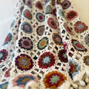 May include: A crochet blanket featuring a repeating pattern of circular motifs in various colors including red, blue, brown, green, and yellow, all joined by an off-white yarn lattice. The edges are finished with small, round, off-white yarn pom-poms.