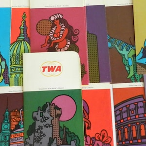 May include: A collection of vintage TWA travel brochures featuring colorful illustrations of various destinations, including a cactus, a castle, a cityscape, and a mountain. The brochures have the TWA logo and the text "Trans World Airlines" on the front.