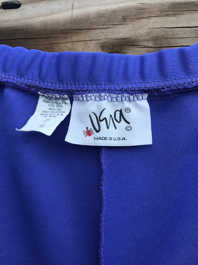 Pode incluir: Uma cal&ccedil;a roxa com uma etiqueta branca que diz "Vera-Tex 100% Poli&eacute;ster Feito nos EUA" e um logotipo de joaninha vermelha com o texto "Usna" e "Feito nos EUA."