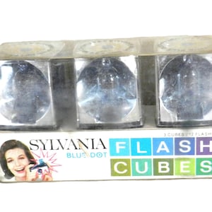 May include: A package of three Sylvania Blue Dot Flash Cubes. The package is white with blue, pink, and purple text that reads "Sylvania Blue Dot Flash Cubes". The package shows a woman using a camera with a flash cube.