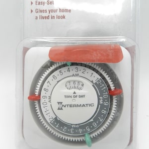 May include: An Intermatic lighting timer in clear plastic packaging. The device features a white circular dial with black numbers for AM and PM, a central time-of-day indicator, and red and green pins for setting on and off times for home lighting.