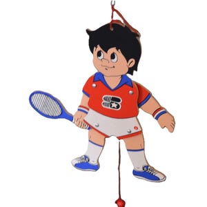 May include: A wooden pull-string toy of a boy playing tennis. He is wearing a red and blue shirt, white shorts, and blue sneakers. He is holding a blue tennis racket. The toy is suspended from a string with a red ball at the end.