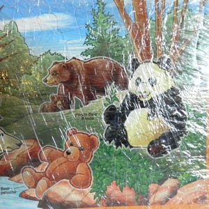 May include: A colorful jigsaw puzzle featuring a forest scene with a brown bear family, a panda bear, and a teddy bear. The puzzle has a blue sky and green trees. Text on the puzzle reads "Panda Bear - Panda" and "Teddy Bear - Ours en peluche".