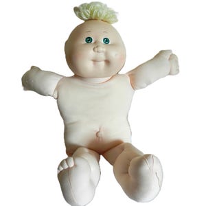 May include: A vintage baby doll with a soft, fabric body and a vinyl head. The doll has a light skin tone, green eyes, and a tuft of light yellow hair. It is wearing a simple, cream-colored outfit and has its arms outstretched.