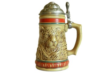 May include: A ceramic beer stein with a lid and handle. The stein features a detailed relief of a leopard's face. The stein is decorated with red, green, and gold accents.