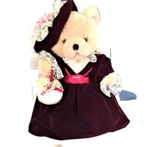 May include: A plush teddy bear wearing a burgundy velvet dress with white lace trim and a pink satin sash. The bear is wearing a white hat with a pink flower and holding a white basket with a pink bow.