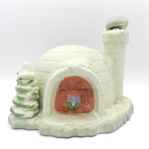 May include: A ceramic igloo figurine with a red door, a chimney, and a small Christmas tree. The igloo is white with a textured surface, and the base is covered in glitter. A festive winter decoration.